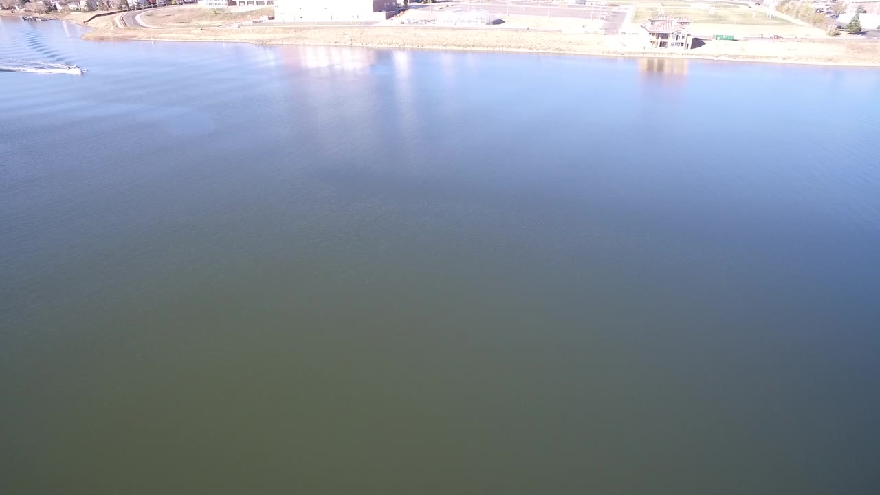 High aerial frame over Hidden Lake, Colorado showing blue green rippled water, a narrow bright shoreline, small structures near the bank, and no visible people.