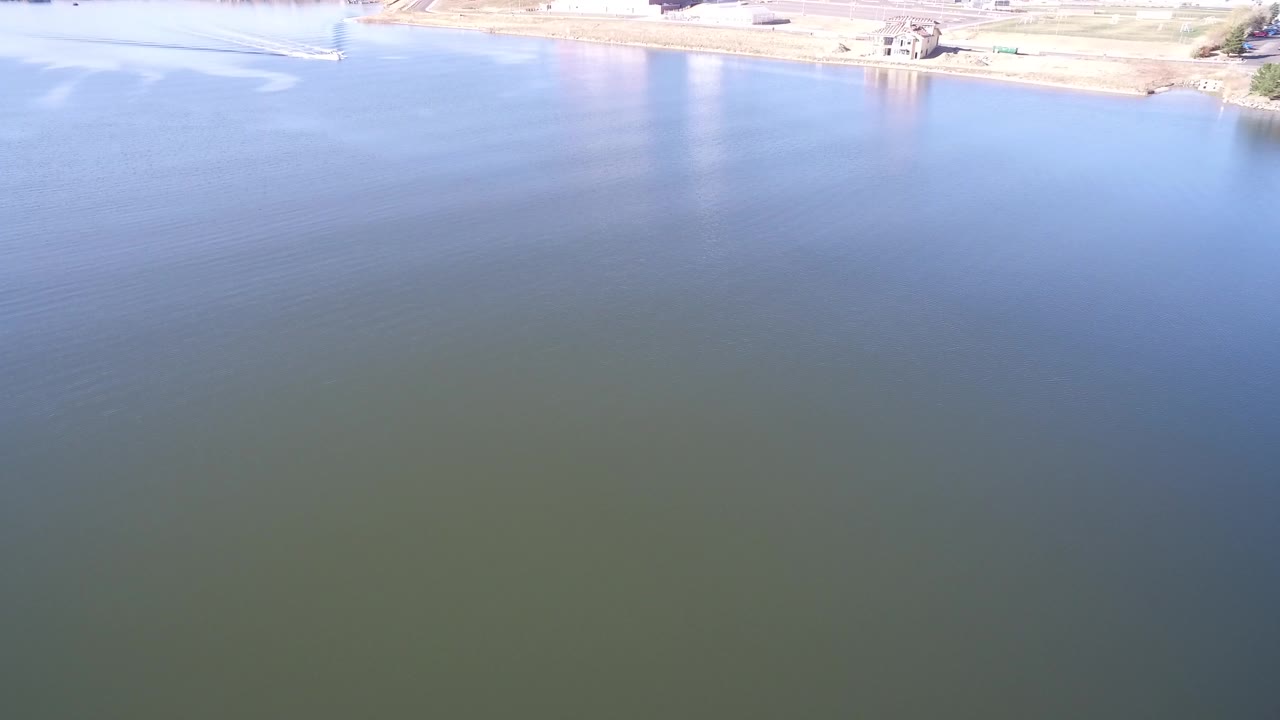 Opening aerial frame over Hidden Lake, Colorado, with blue-green rippled water filling the view, a bright shoreline along the top edge, and no visible people.