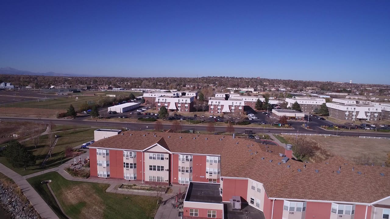 Second aerial frame in Hidden Lake, Colorado with the tan-roofed building lower in frame, open grounds and roads across the middle, distant low buildings, tree line, mountains, and no visible people.