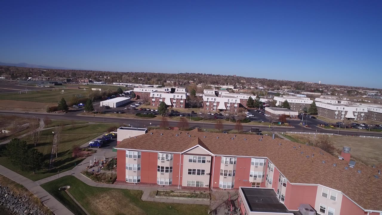 Aerial frame in Hidden Lake, Colorado showing the pink building closer across the lower foreground, with parked cars, a road, white low-rise buildings, and no visible people.