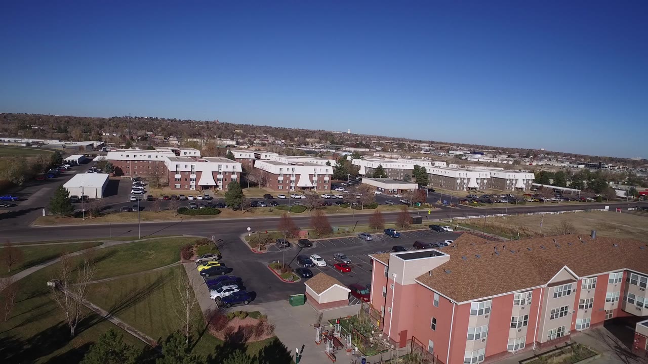 Aerial view over apartment buildings, lawns, parking lots, and a road under a clear sky.