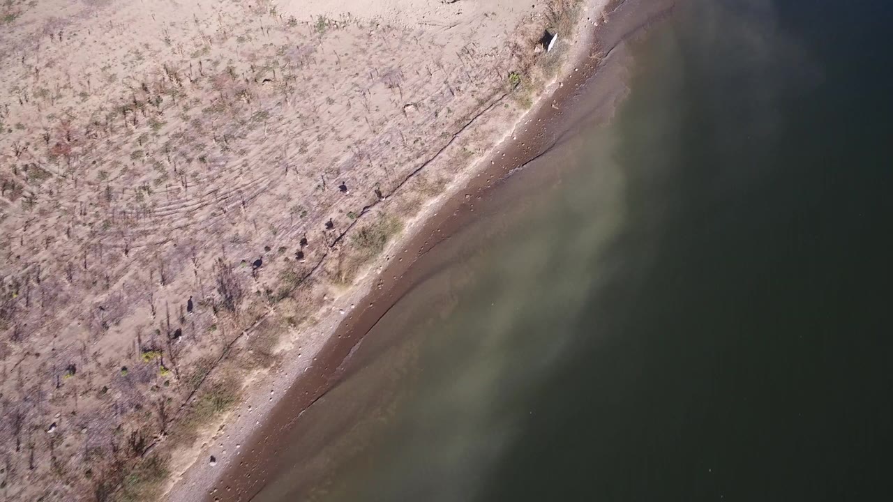 High aerial view over Hidden Lake, Colorado, with the shoreline shifting across the frame, darker water at right, pale exposed ground at left, and no visible people.