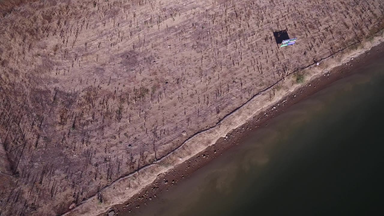 Overhead frame in Hidden Lake, Colorado showing the lake edge running diagonally below brown brush, with a small dark structure near the upper shoreline and no visible people.
