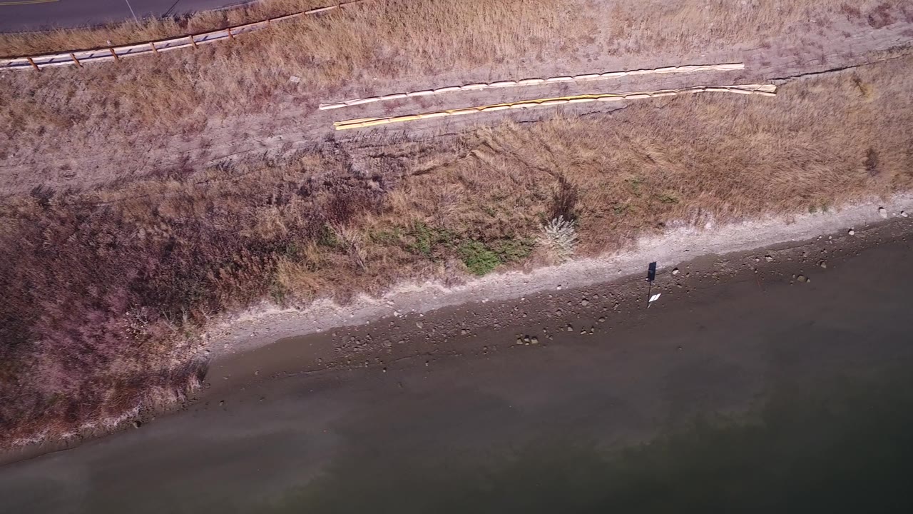Aerial frame over Hidden Lake, Colorado with a curved gravel shore, dark green water, brown brush, a dirt track, and no visible people.