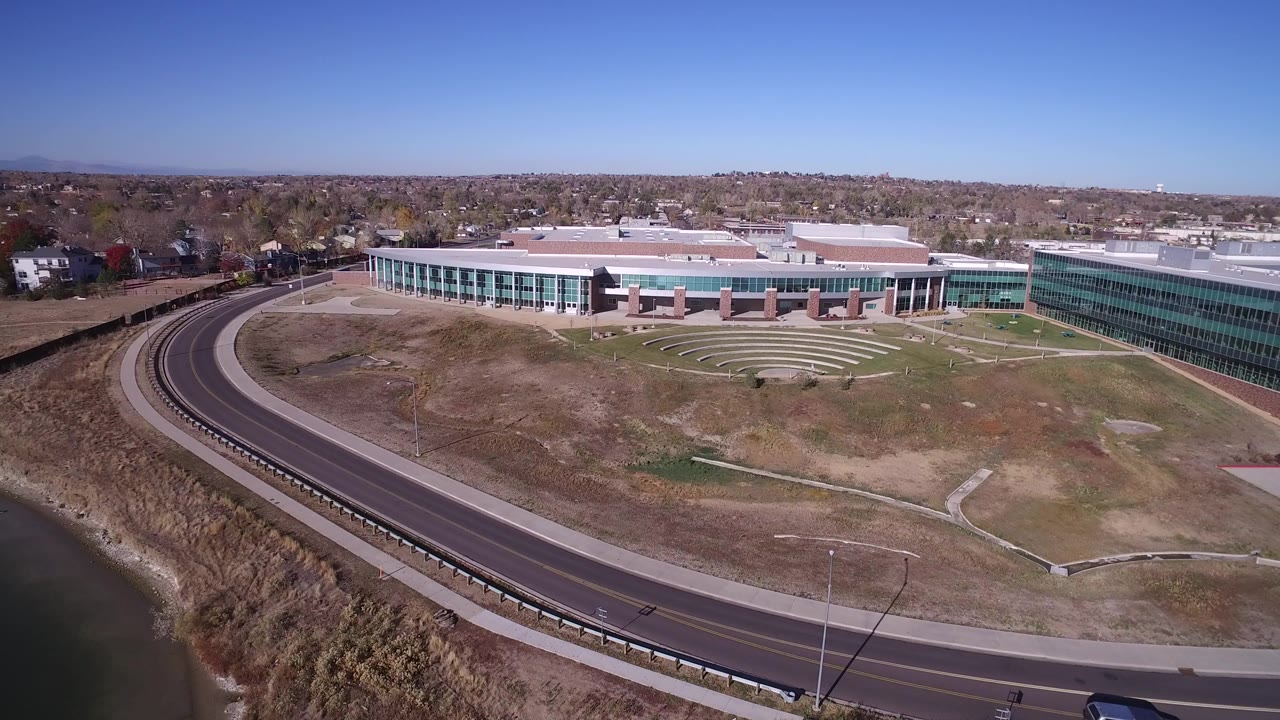 High aerial frame over Hidden Lake, Colorado showing a large modern building, curved road, shoreline water, dry open ground, distant houses, blue sky, and no visible people.