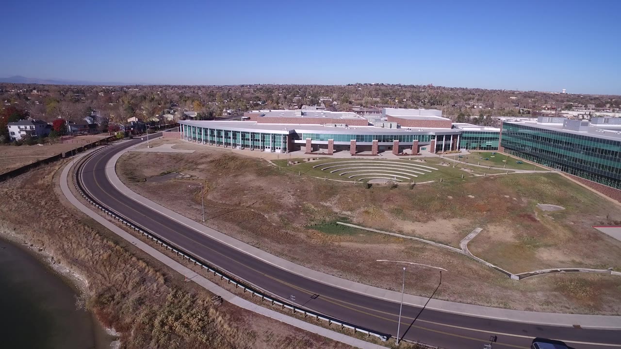 Aerial frame in Hidden Lake, Colorado with more of the curved roadway visible below the large glass building, lake water at the lower edge, distant neighborhoods, and no visible people.