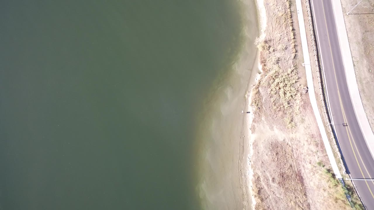Overhead view of green lake water beside sandy shore and a paved road.