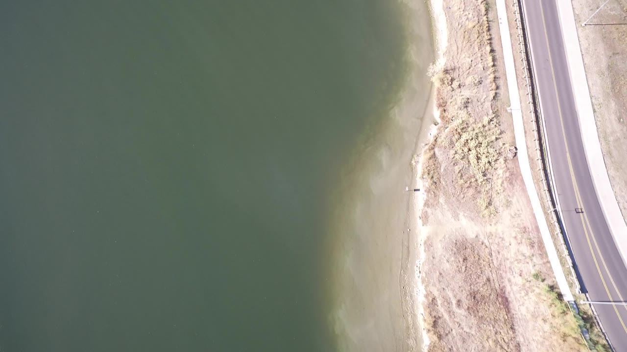 At one second, the Hidden Lake Colorado shoreline remains centered between muted green water and the paved road, with bright exposed sand and no visible people.