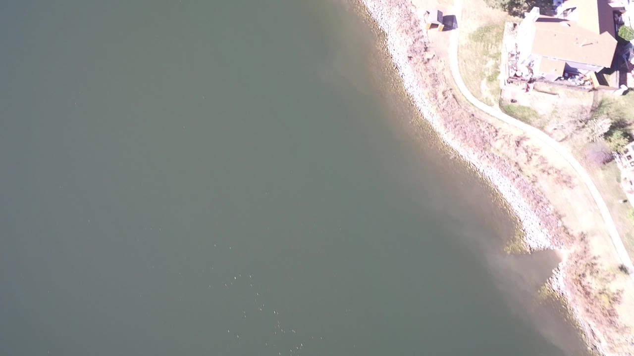 Aerial frame at Hidden Lake, Colorado with still green water on the left, a pale curved shore through the center, and houses clipped along the upper right, with no visible people.