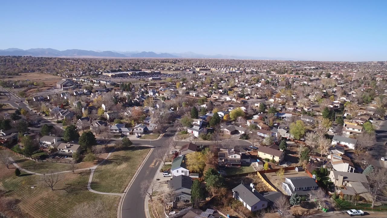 Opening aerial frame over Hidden Lake, Colorado, with curved neighborhood streets, private homes, open grass, scattered trees, a clear blue sky, and no visible people.