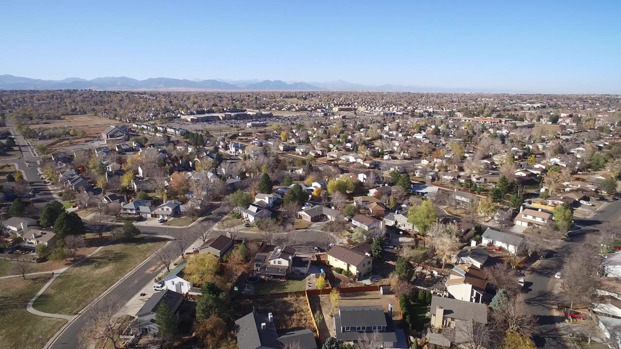 Opening aerial frame over Hidden Lake, Colorado, showing private homes, curving roads, scattered trees, brown lawns, distant mountains, clear blue sky, and no visible people.