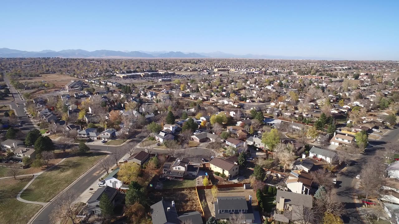 One second into the Hidden Lake, Colorado aerial, rooftops and residential streets fill the foreground while a wide band of suburban blocks stretches toward distant mountains, with no visible people.