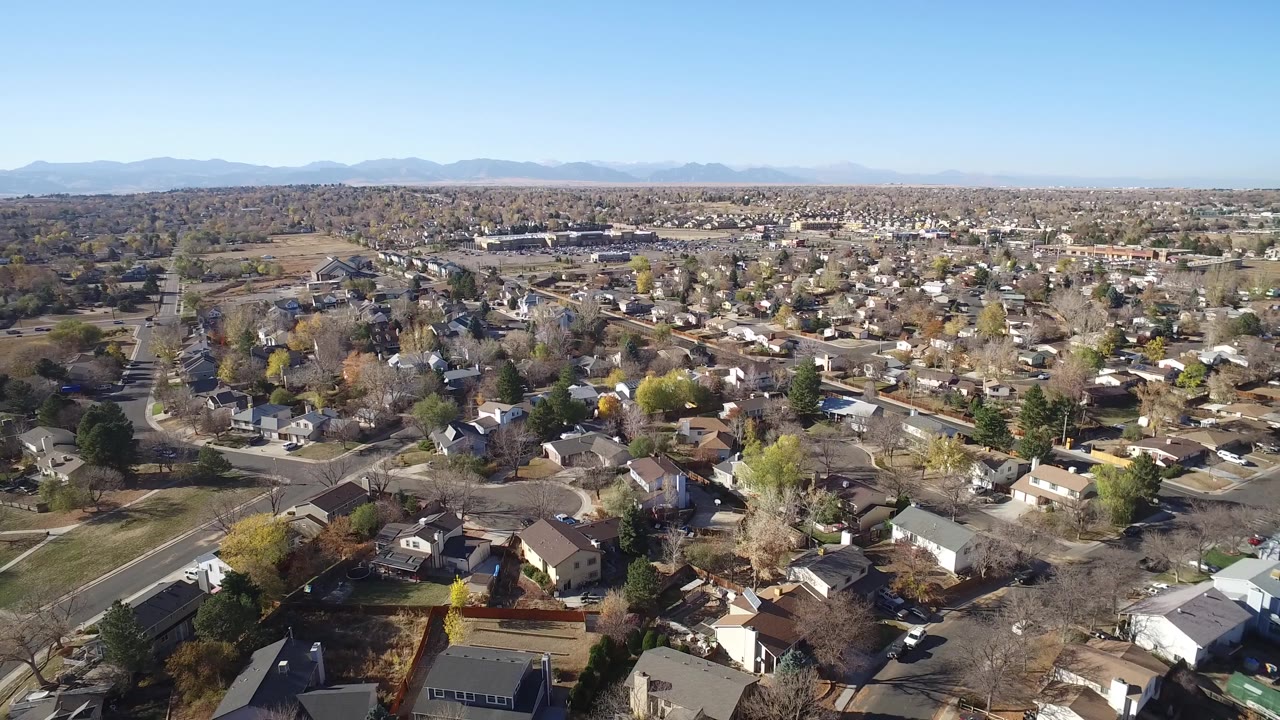 Wide aerial view of suburban homes, streets, trees, mountains, and blue sky.