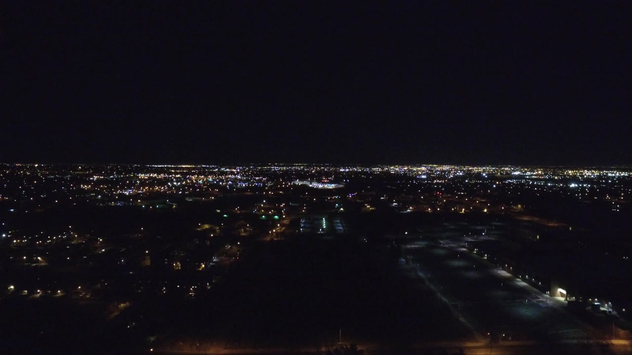 Denver night aerial at 0:01 showing a broad band of white and amber lights, a dim road grid, dark open areas below, and no visible people.