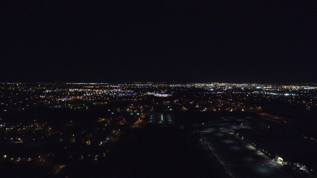 Aerial nighttime view over Denver with clusters of white and amber lights along the horizon, dark open foreground areas, and no visible people.