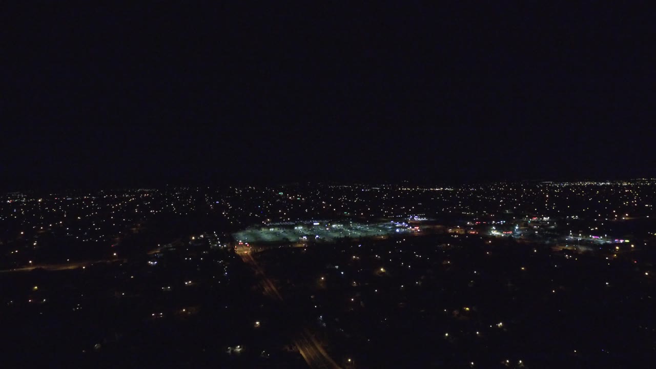 Elevated Denver night frame with a broad band of urban lights across the horizon, dim foreground blocks, and no visible people.