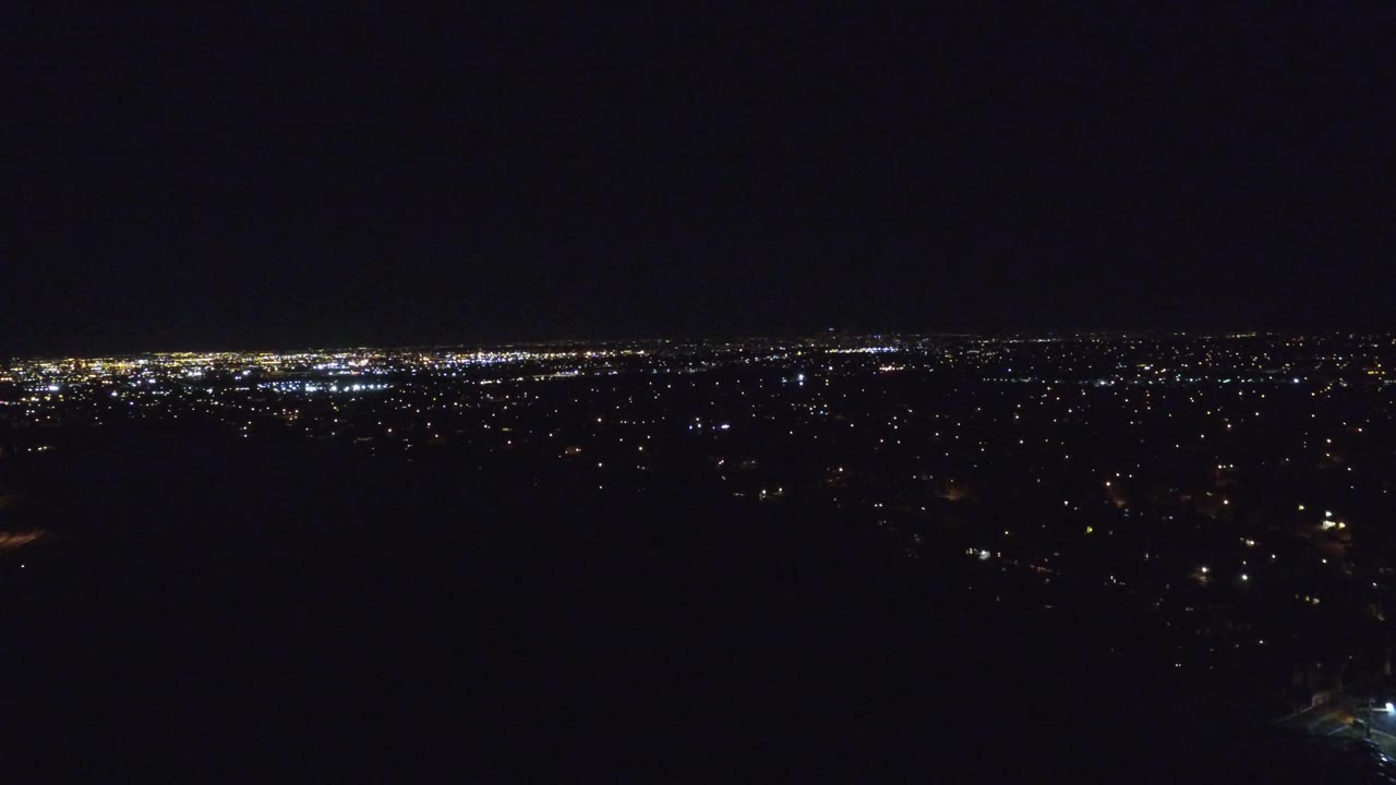 Aerial night frame of Denver, Colorado, with clustered white and amber lights across the horizon and sparse lights in the dark foreground, no visible people.