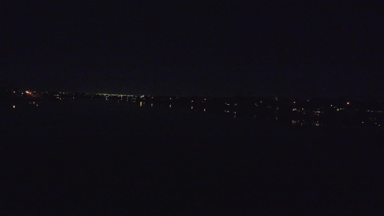 A dark Denver lakeside view shows scattered points of light reflected on the water, with the horizon slightly clearer and no visible people.