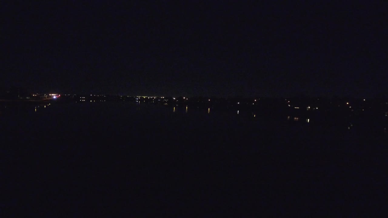 Dark Denver water view one second in, with faint reflected lights spread along the shoreline and no visible people.