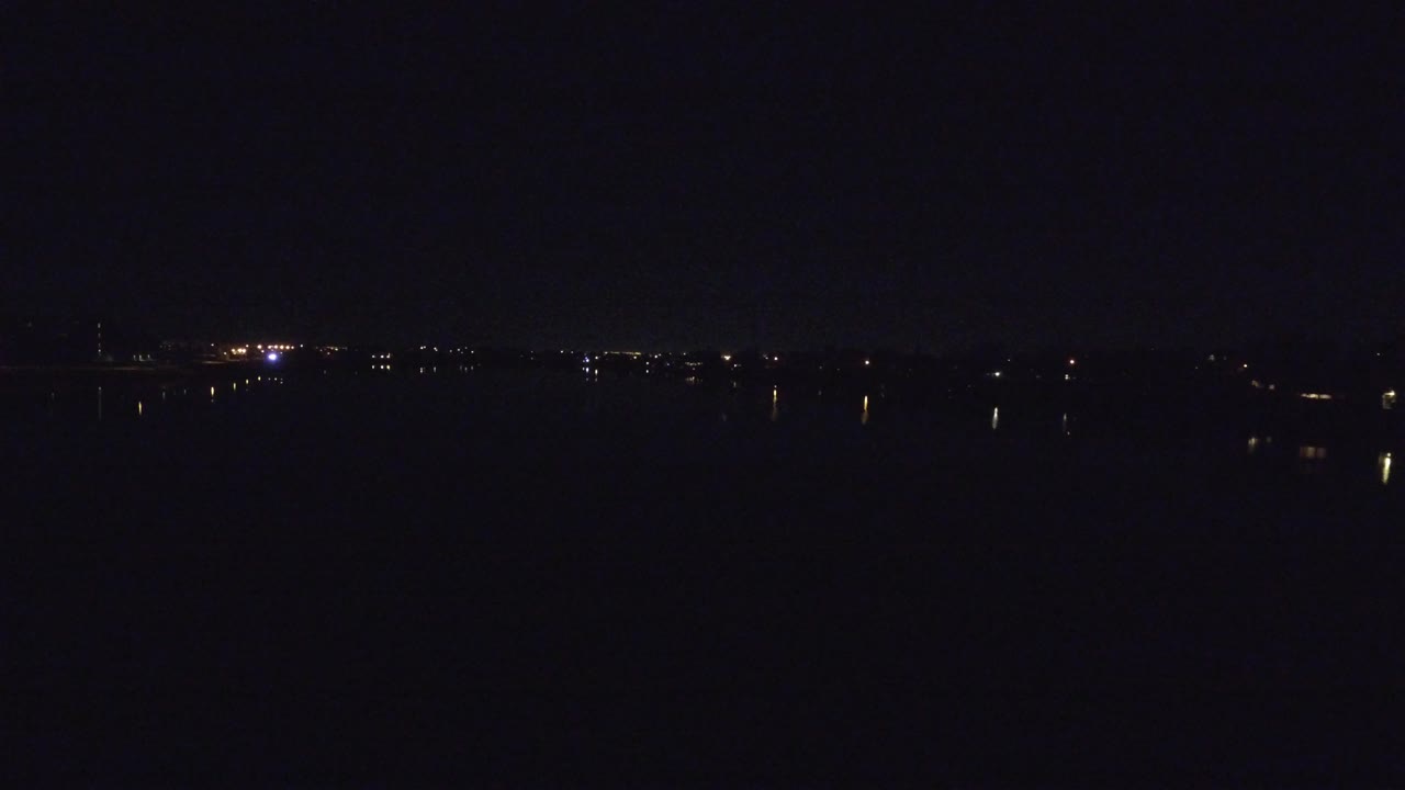 A very dark Denver lakeside view at night, with scattered shoreline lights and faint reflections on the water, no visible people.