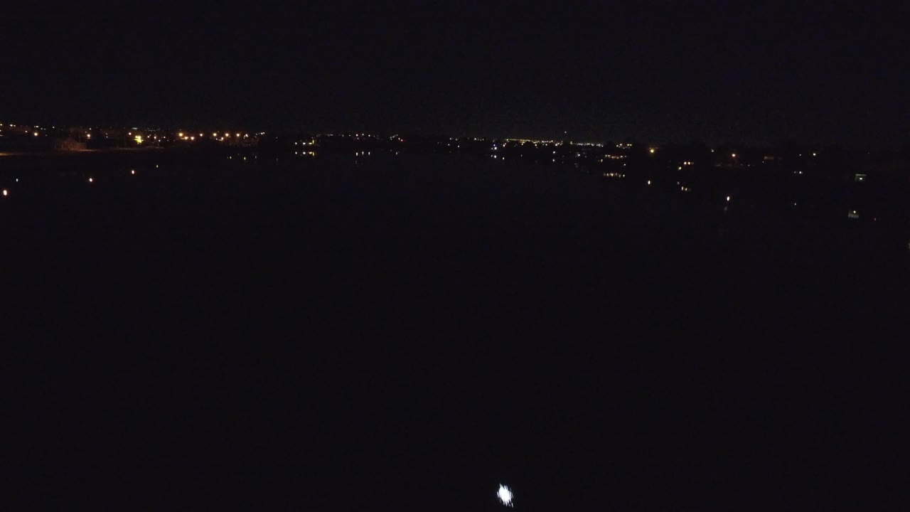 Bright vessel light on a dark river with a band of distant lights behind it.