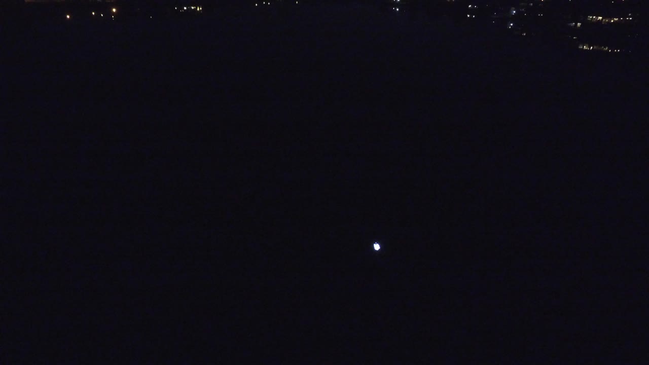 At 0:00 in Denver, Colorado, a dark lake fills the frame with a small bright moon reflection near the center and faint distant lights along the top edge, no visible people.