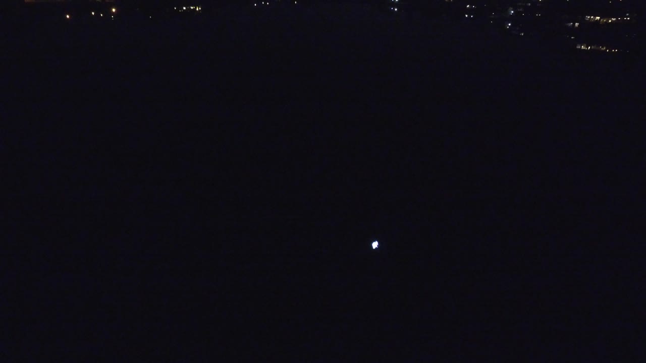 At 0:01 in Denver, Colorado, the bright reflection remains centered over a dark lake while small shoreline lights sit high in the frame, no visible people.