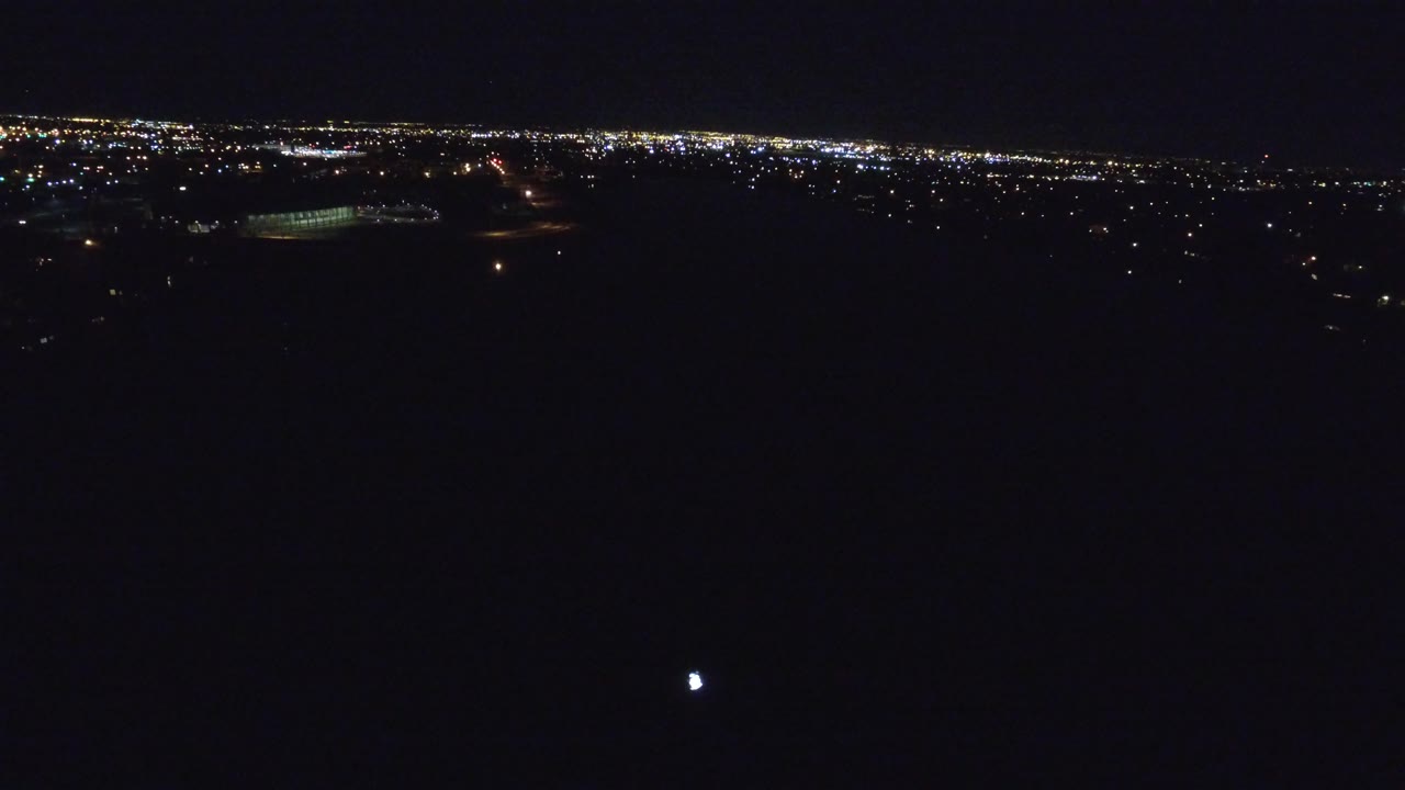 A high nighttime view over Denver shows scattered lights below, brighter urban light along the horizon, and no visible people.