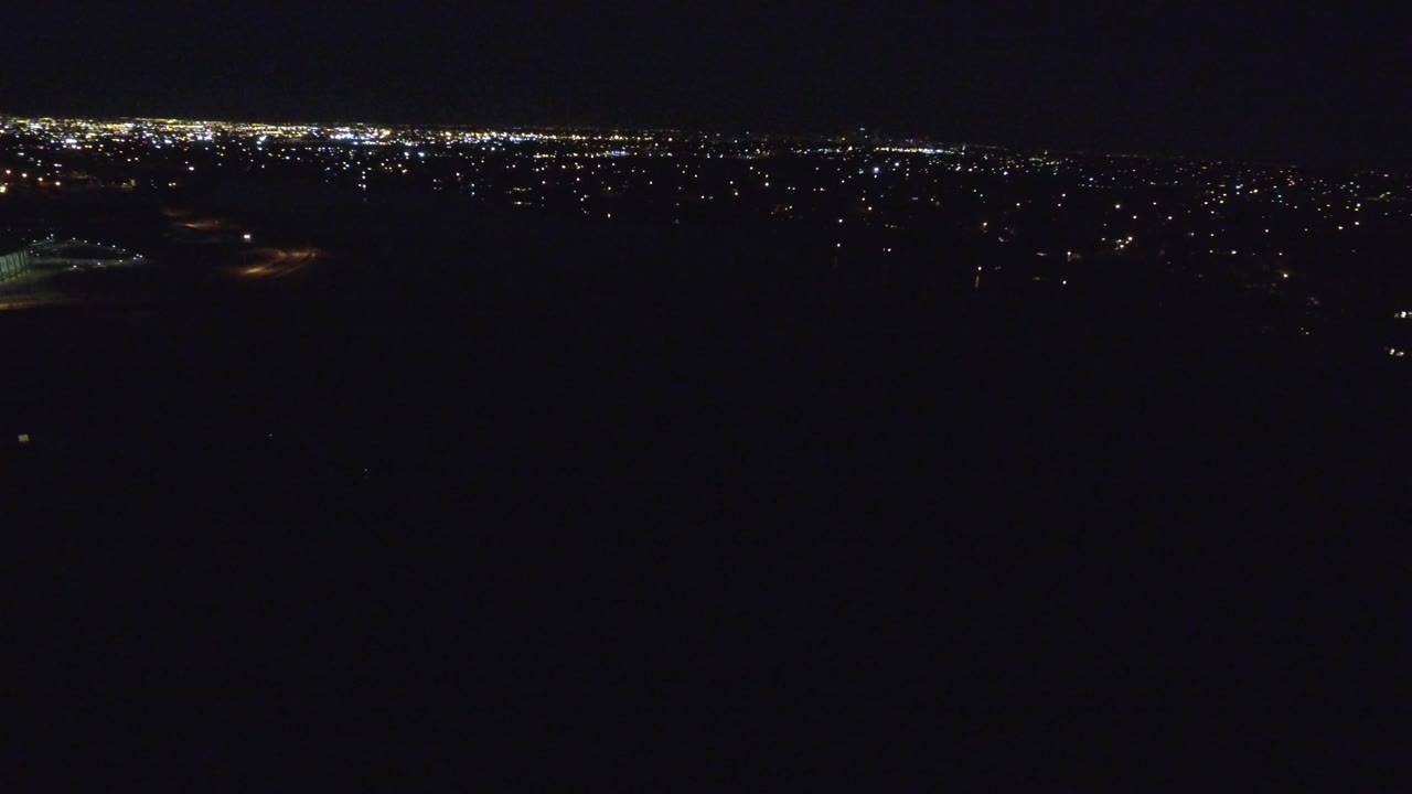 Second frame of the Denver night aerial, showing a bright band of distant city lights across the upper frame and broad black foreground with no visible people.