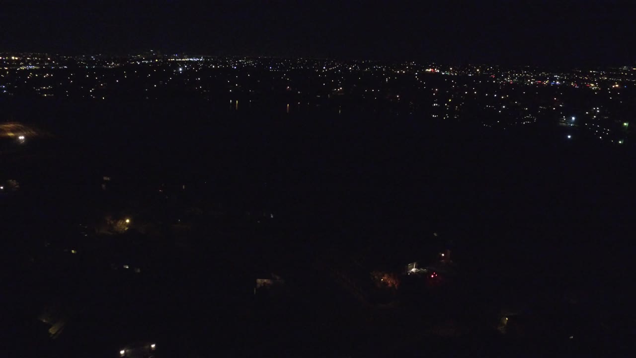 Denver nighttime aerial frame showing sparse light clusters below and a brighter strip of city lights across the upper background, with no visible people.