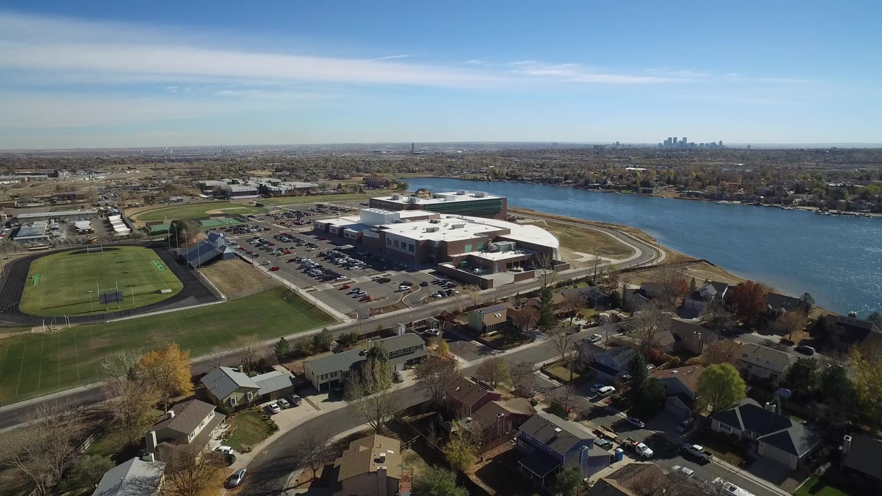Opening aerial frame over Westminster, Colorado with rooftops in the foreground, a green sports field at left, a large building complex near the river, distant skyline, and no visible people.