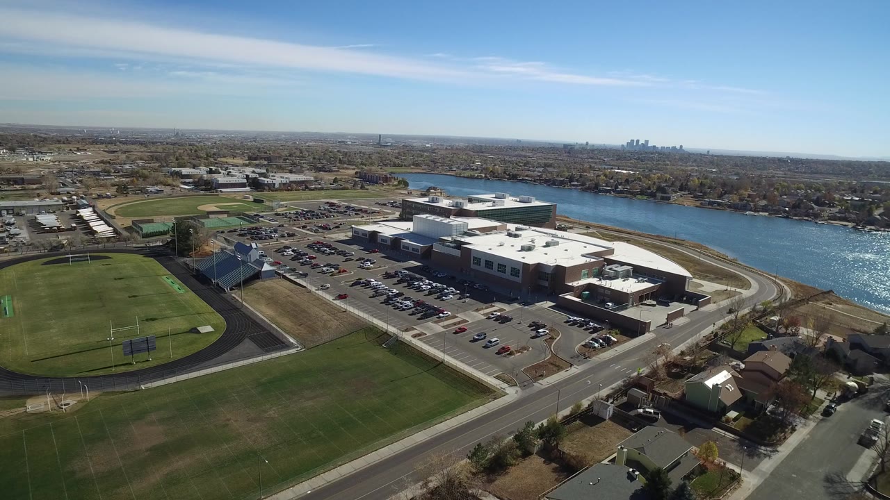 Wide aerial frame over Westminster, Colorado showing a track field at left, a large low building and parking lot near the water, suburban blocks beyond, and no visible people.
