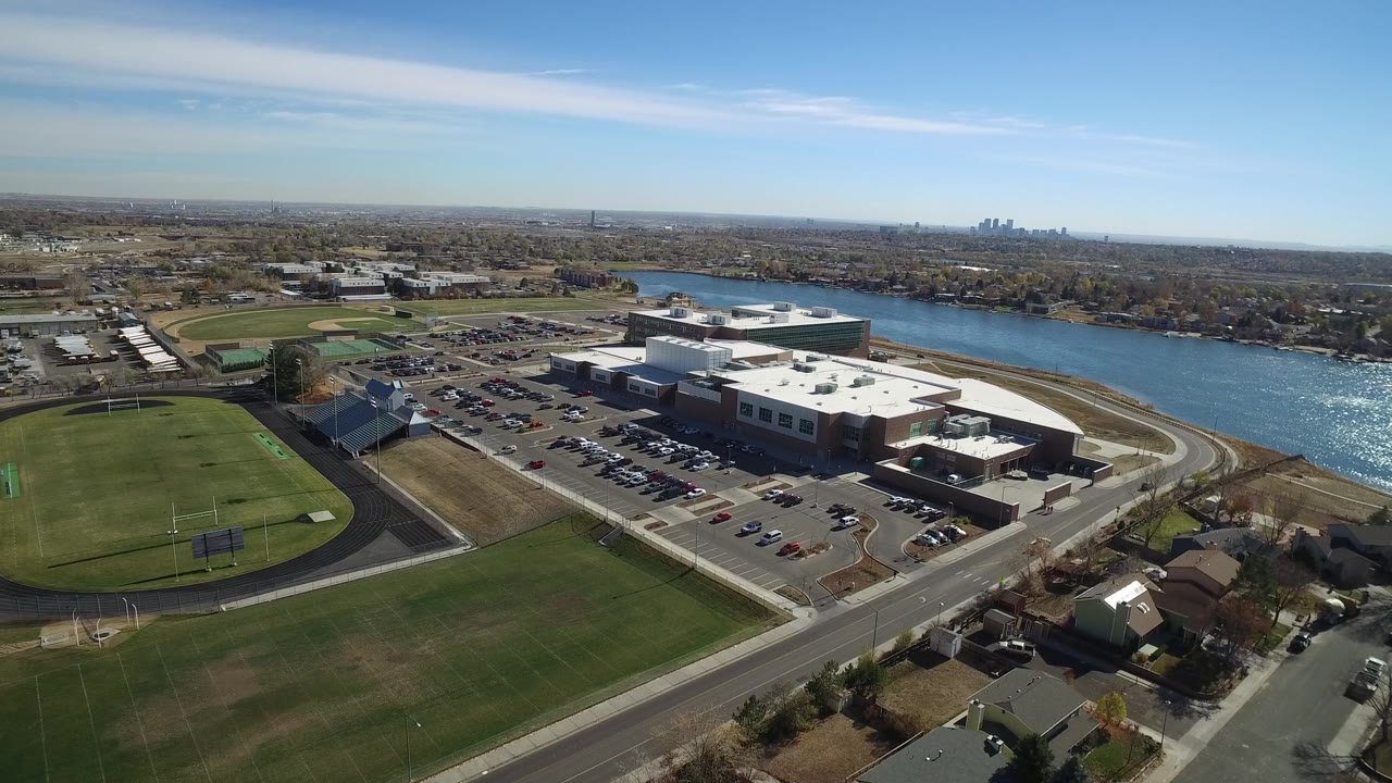 Second aerial frame in Westminster, Colorado with the athletic fields lower left, the large building and parking lot centered, bright water behind it, and no visible people.