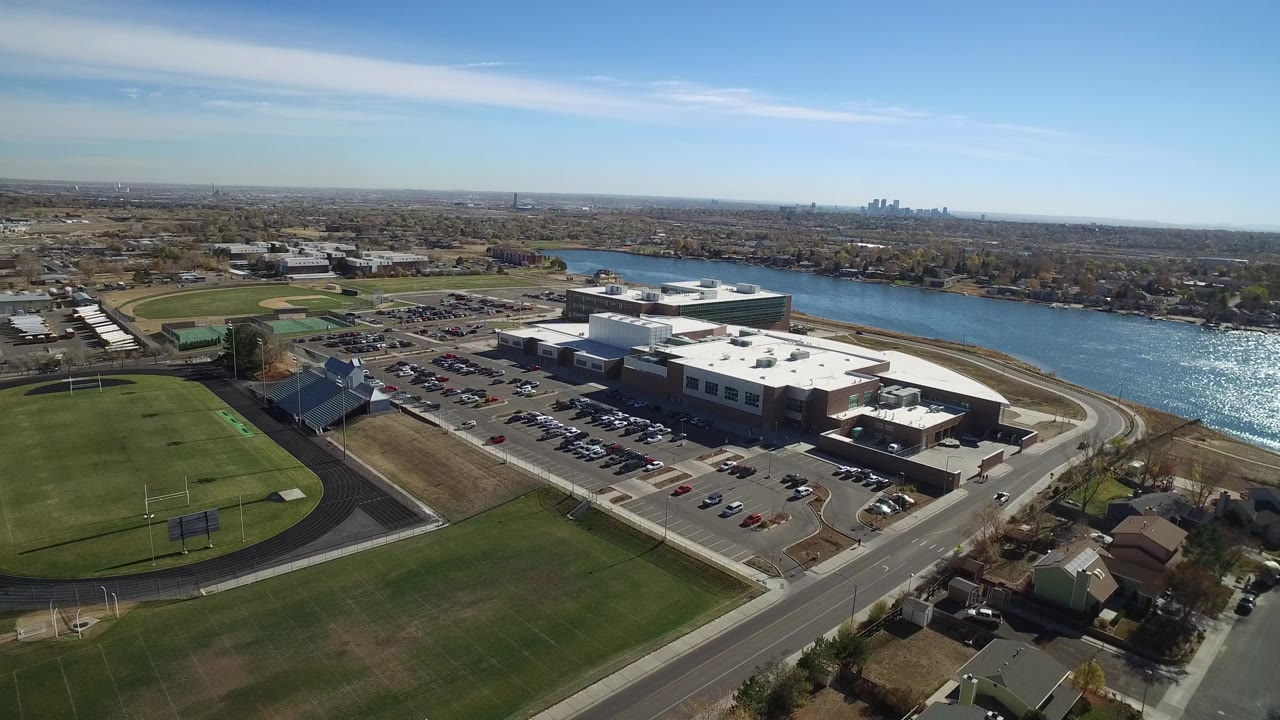 High aerial view of sports fields, parking, a large building, water, suburbs, and skyline.