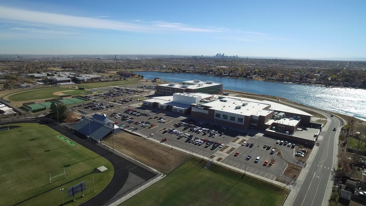 Aerial frame over Westminster, Colorado showing athletic fields in the foreground, a mall and parking lots beside a sunlit lake, distant skyline on the horizon, and no visible people.