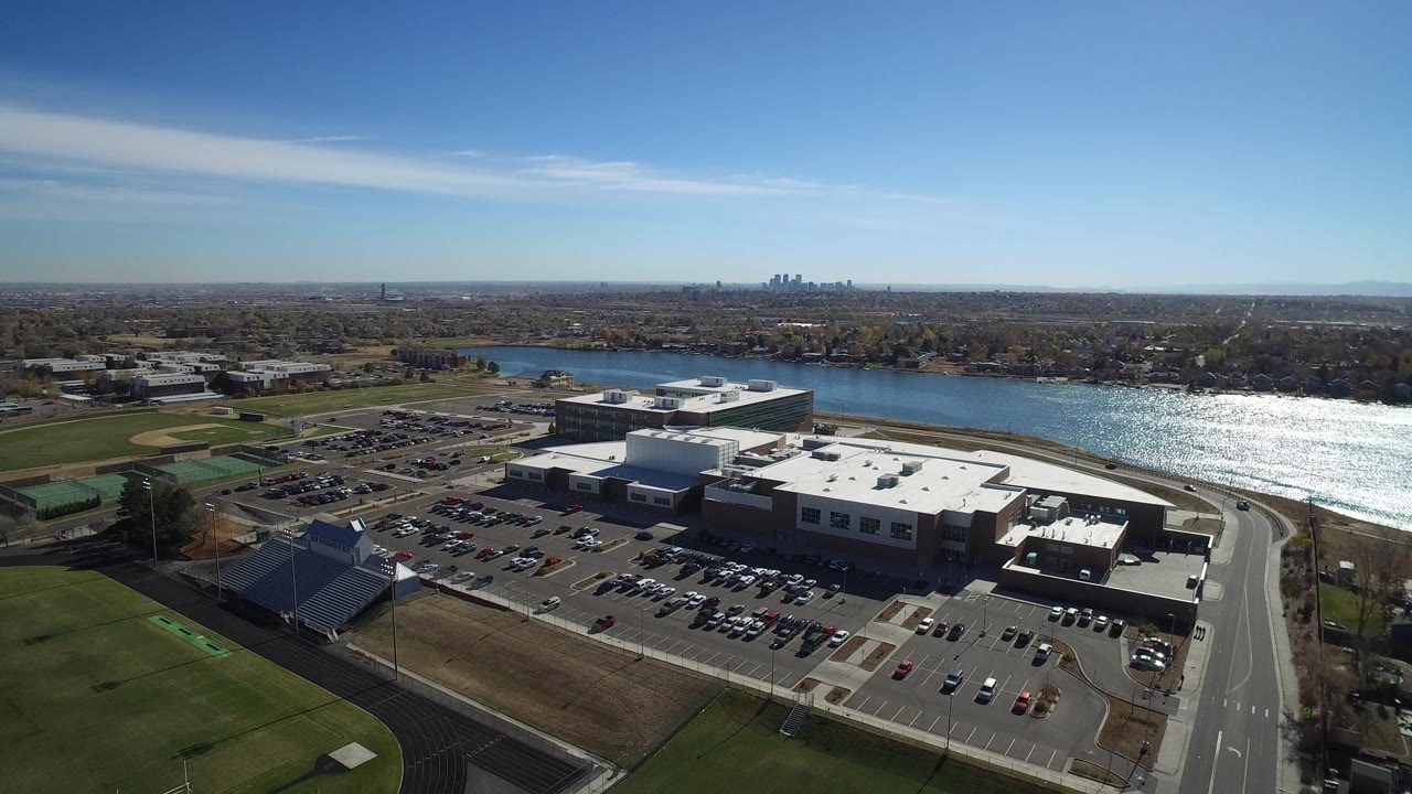 Wide aerial view in Westminster, Colorado looking over sports fields, parking lots, roads, a bright lake, distant skyline, and no visible people.