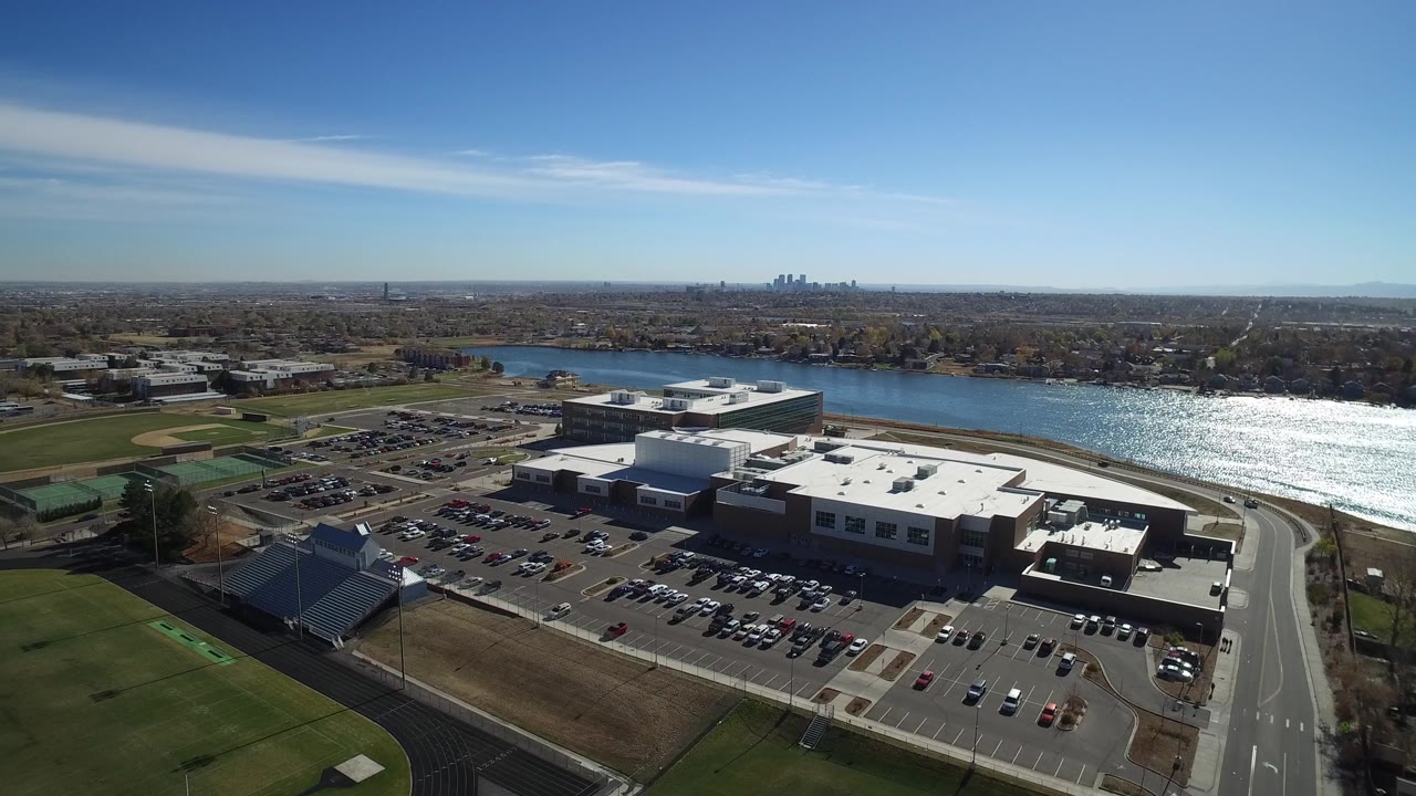 Aerial city edge view with lake, parking lots, sports fields, buildings, and a clear horizon.