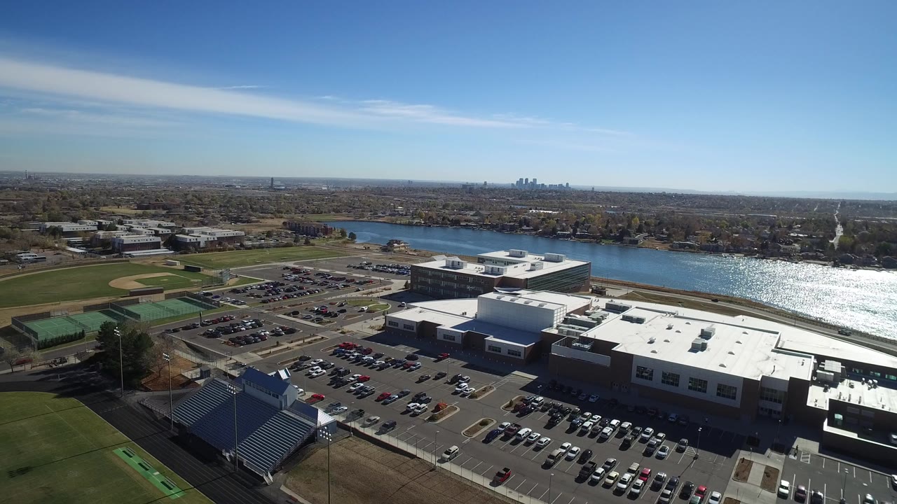 At 0:00, a high aerial view over Westminster, Colorado shows white-roofed buildings, parked cars, ball fields, a river, distant skyline, and no visible people.