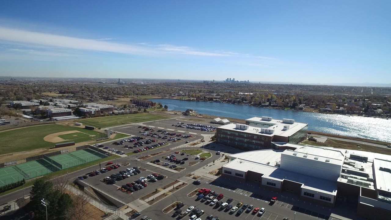 Aerial view of tennis courts, parking lots, buildings, fields, water, and distant skyline.