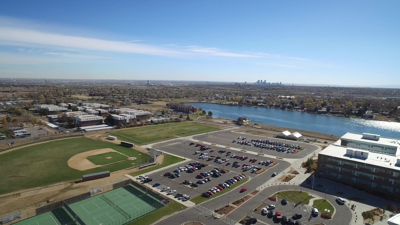 High aerial frame in Westminster, Colorado with baseball diamonds on the left, a large parking lot and building to the right, water beyond, and no visible people.