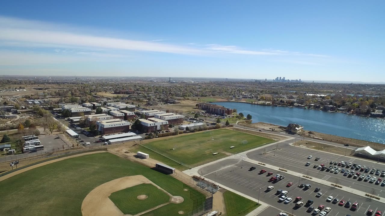 Aerial view of baseball field, parking lot, buildings, water, and distant skyline under blue sky.