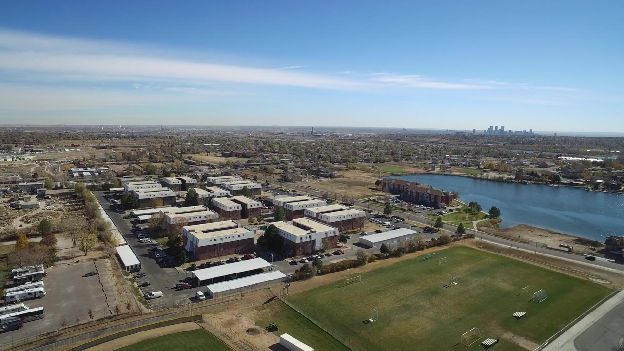 Opening aerial view in Westminster, Colorado, showing low-rise commercial buildings beside a road, a green field, a blue lake, and a distant skyline with no visible people.