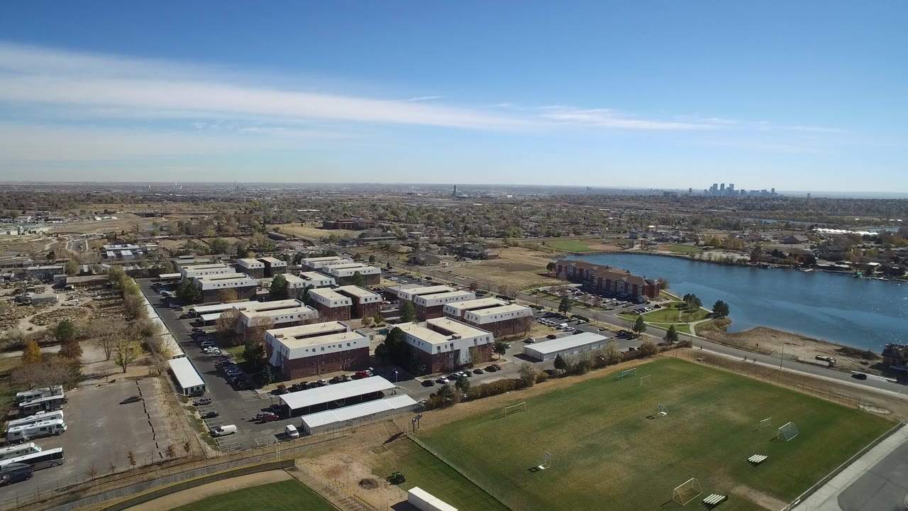 Aerial frame in Westminster, Colorado, with flat-roofed office buildings and parking areas in the foreground, the lake to the right, and a distant skyline under clear sky with no visible people.