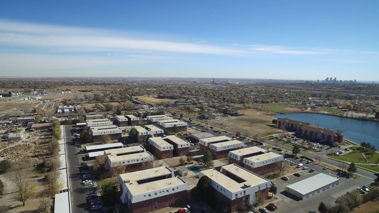 Opening aerial frame over Westminster, Colorado showing low-rise residential buildings, parking lots, a lake to the right, distant city towers, and no visible people.