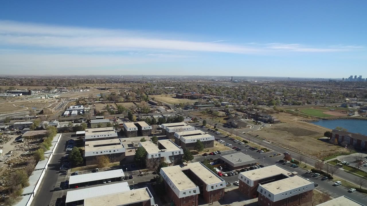 Aerial view over low-rise buildings, roads, fields, and distant skyline under blue sky.