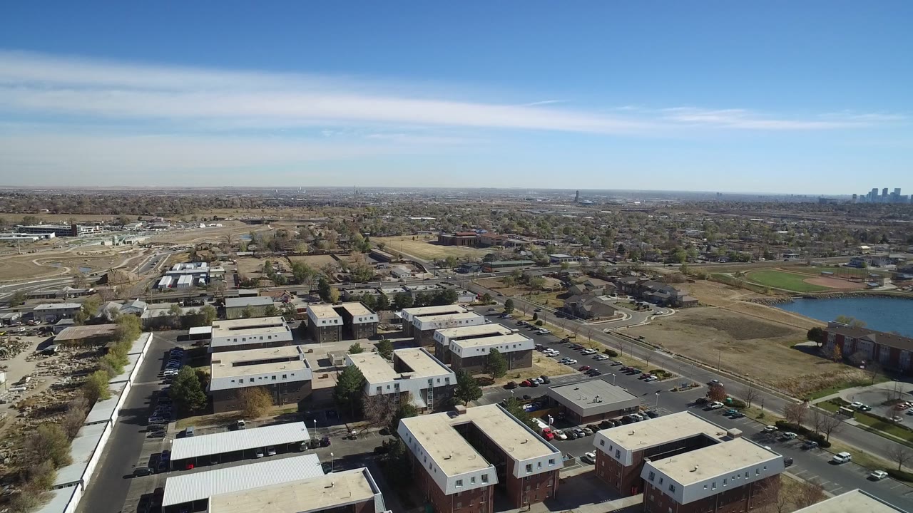 A second aerial view of Westminster, Colorado, keeps the commercial rooftops low in frame while roads, brown open lots, tree-lined neighborhoods, and distant city towers stretch to the horizon with no visible people.