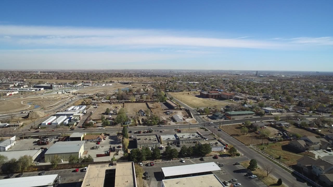 Aerial view over suburban roads, low buildings, open lots, and distant neighborhoods under blue sky.