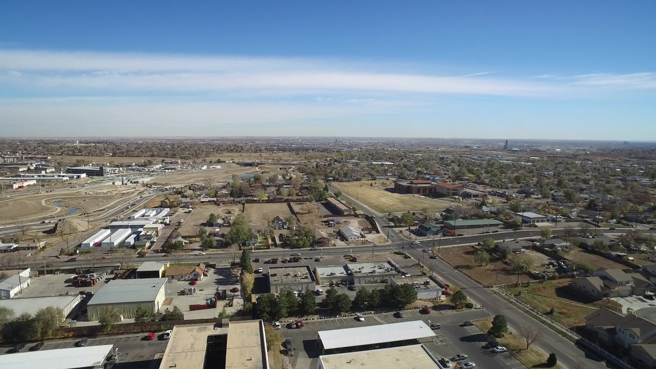 Aerial view over Westminster, Colorado with a diagonal road, warehouse-like buildings, parking areas, tan open ground, trees, distant residential blocks, and no visible people.