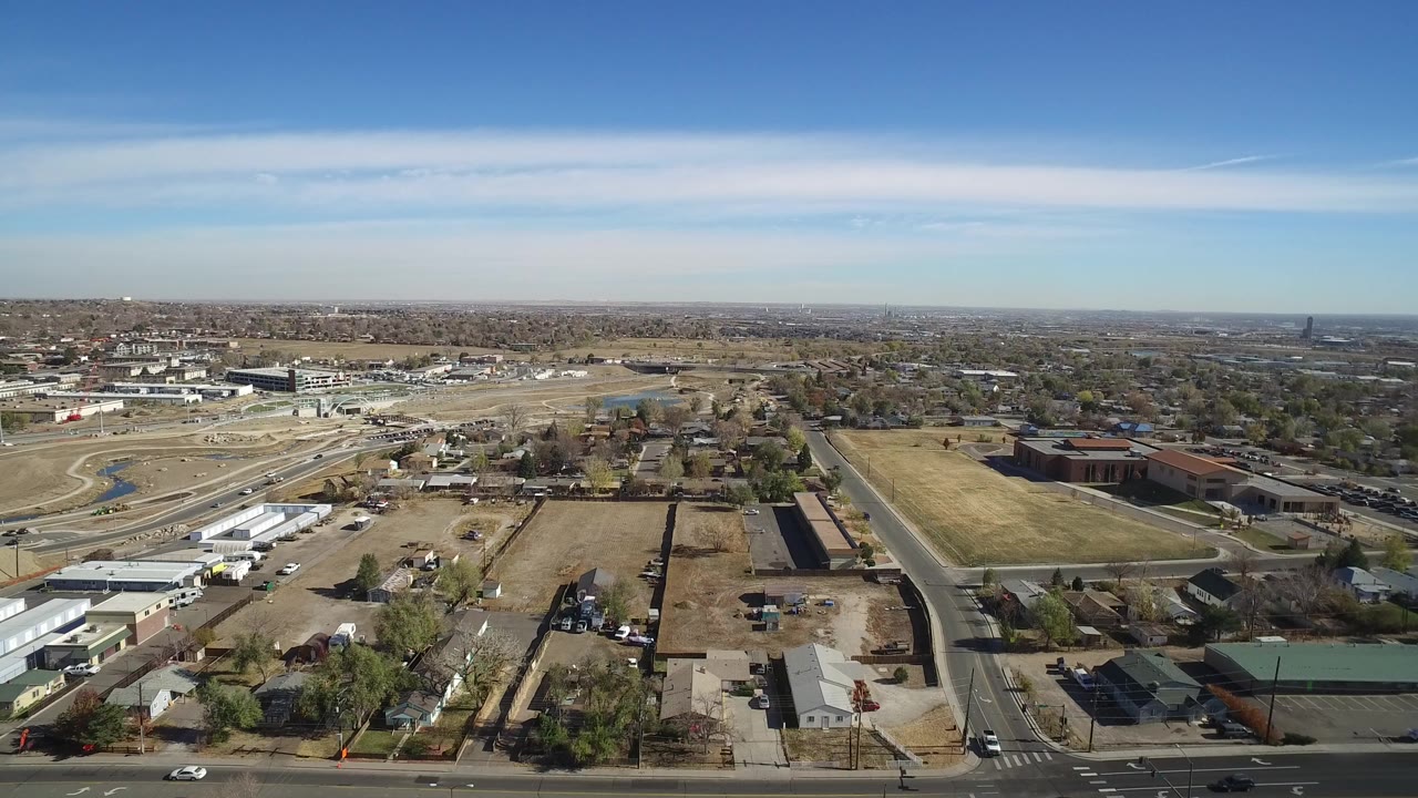 Opening aerial frame over Westminster, Colorado, showing a broad suburban grid with roads, low buildings, homes, open lots, a distant horizon, and no visible people.