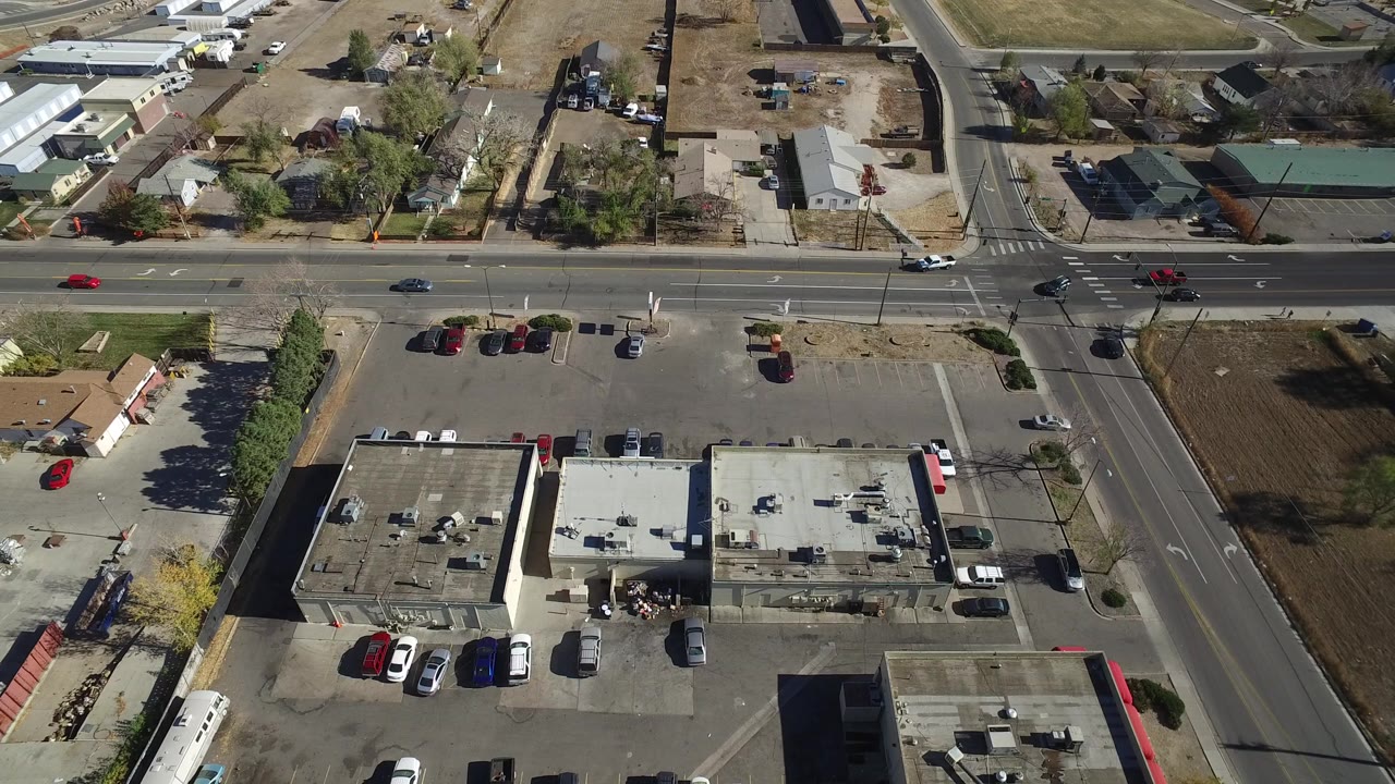 Overhead view of a suburban intersection with rooftops, parking lots, cars, homes, and dry grass lots.